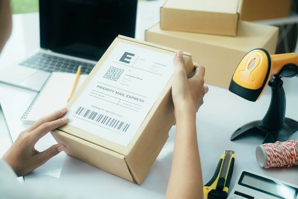 primer-plano-joven-propietaria-negocio-linea-sentada-escritorio-computadora-portatil-cajas-poniendo-etiqueta-envio-cajas-carton-o-paquete-pedido-cliente-preparandose-envio-entrega_1421-9158(1).webp